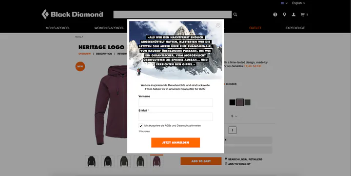 Black Diamond product page for a hoodie, dimmed behind a sign-up pop-up. The pop-up shows two climbers on snow below a granite peak, with a quoted passage in German about climbing the last 300 metres to the summit, then a line promising more travel stories and photographs in the newsletter, fields for first name and email, a consent checkbox, and an orange “Jetzt anmelden” button.